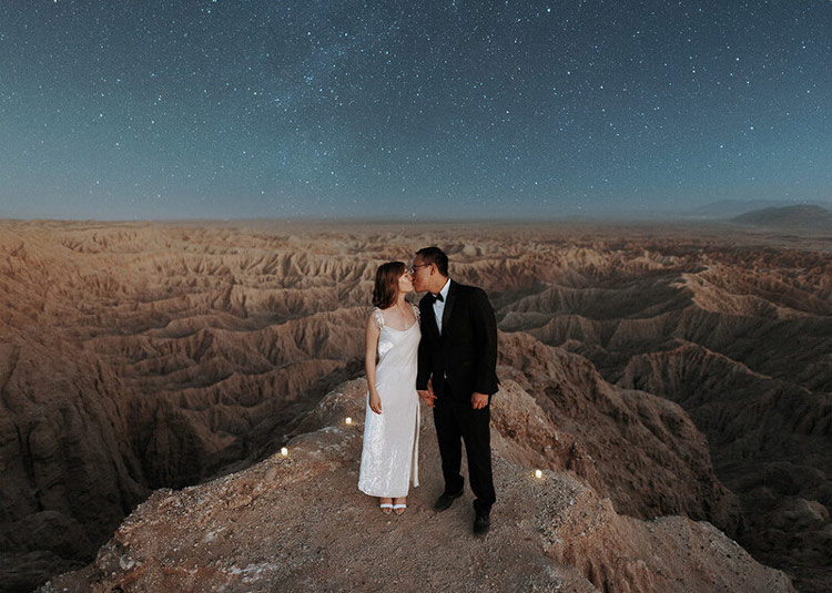 An engaged couple in wedding outfits kissing under the stars with a canyon at Anza Borrego in the background.