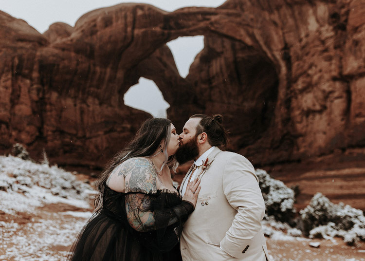 A plus size wedding couple with the bride wearing a black dress kissing at Double Arch in Arches National Park.