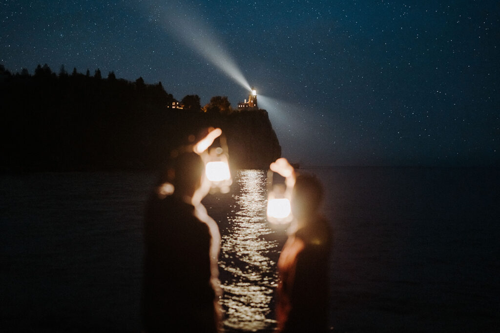 Two men holding lanterns under the stars with Split Rock Lighthouse and Lake Superior in the background.