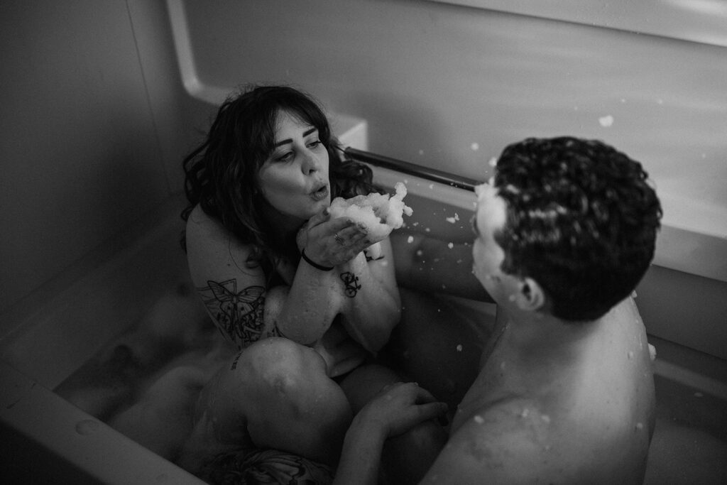 A couples boudoir session with a nude couple sitting in a bathrub. The woman is blowing bubbles at the man.