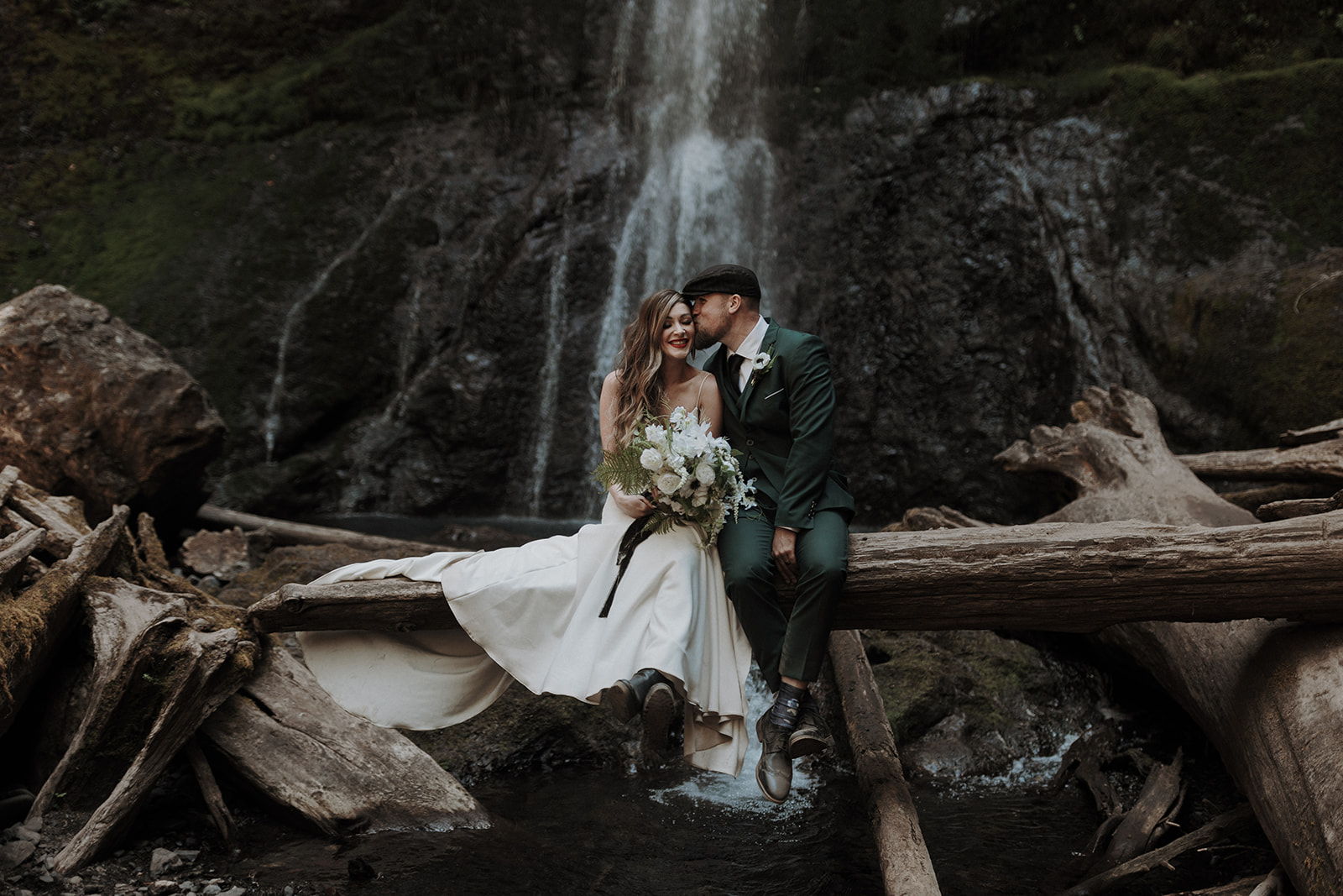 A wedding couple with the groom in a green suit sitting on a log at Marymere Falls in Washington in Olympic National Park.