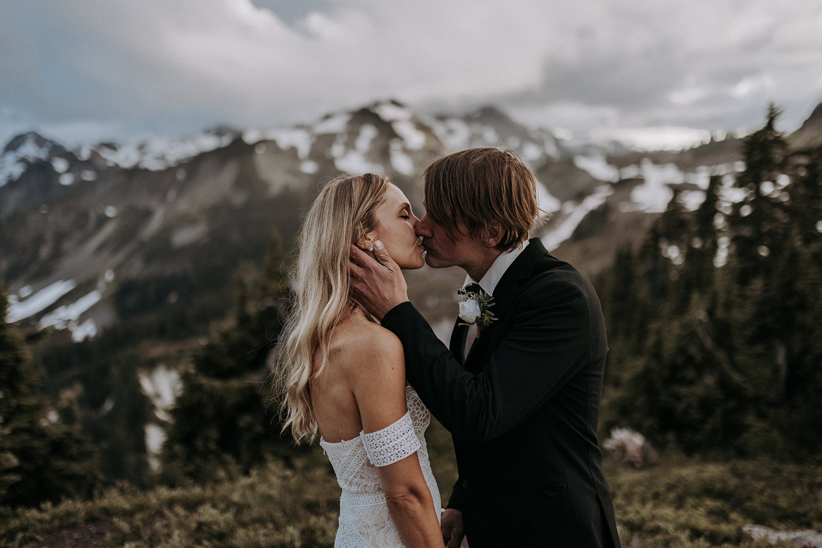 A bride and groom kissing on a mountain with patches of snow in the background in North Cascades National Park.