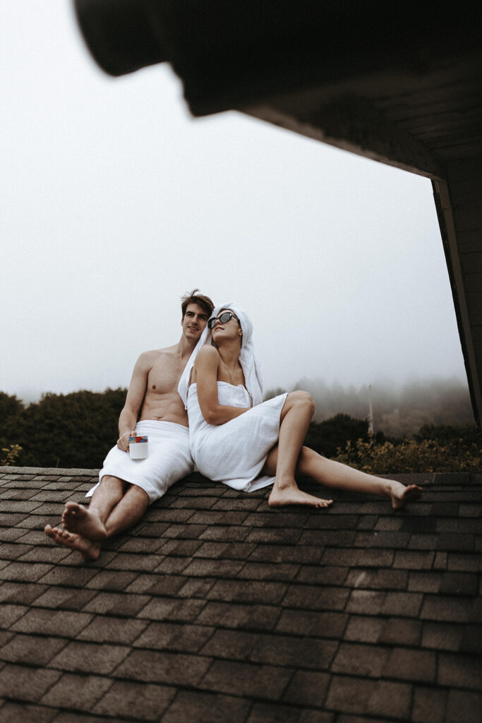 A woman in a towel with sunglasses posing next to her partner who is also in a towel and is holding a mug during their couples boudoir photo session