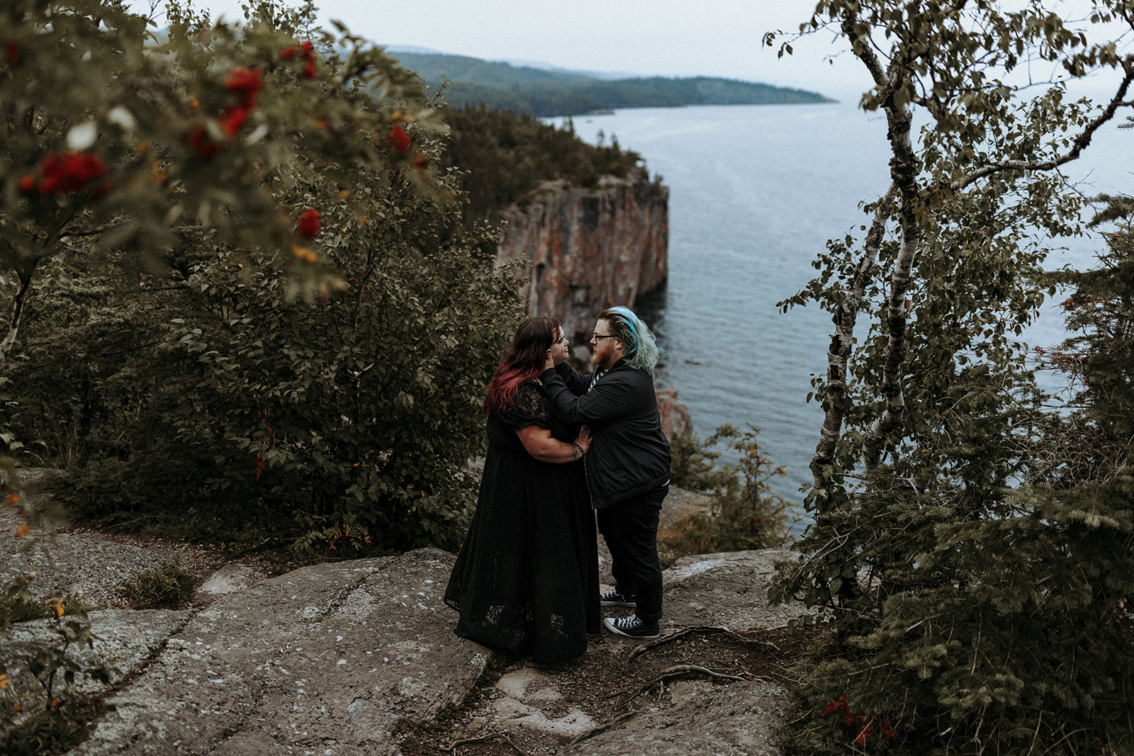 A gothic plus size wedding couple in all black clothes standing near a cliff with red roses in the foreground.