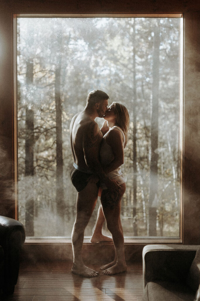 A couples boudoir session with a man and woman kissing in their underwear with a window and the forest behind them.