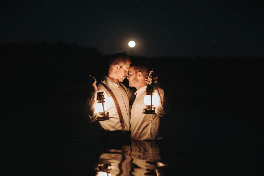 Two grooms in the water under the night sky holding lanterns. 
