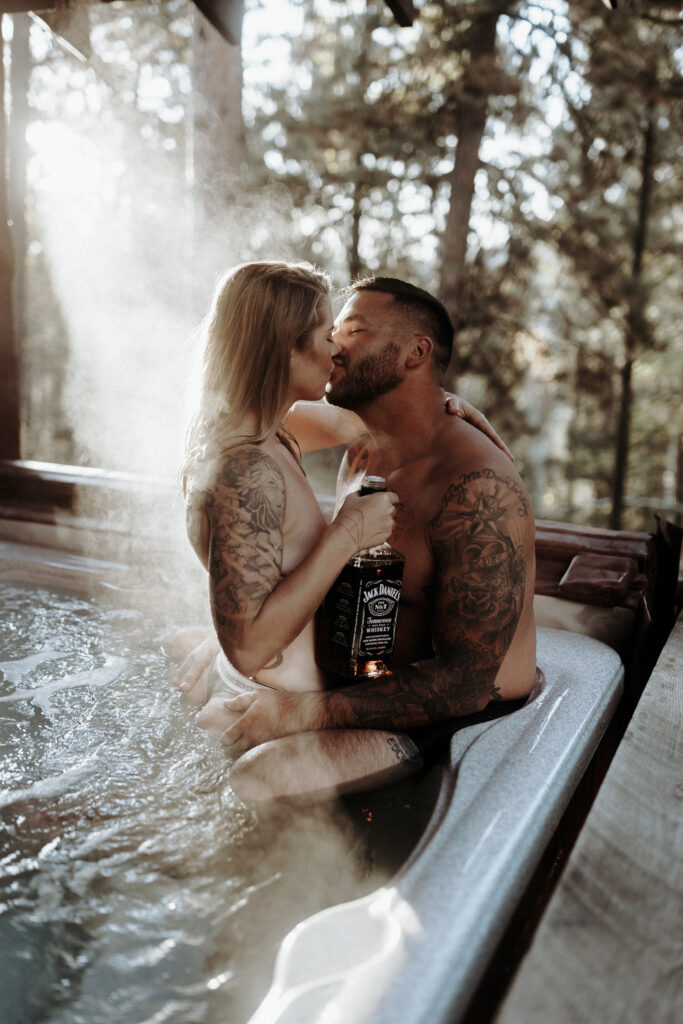 A a couples boudoir session with man and woman kissing shirtless in a hot tub with a bottle of whiskey.