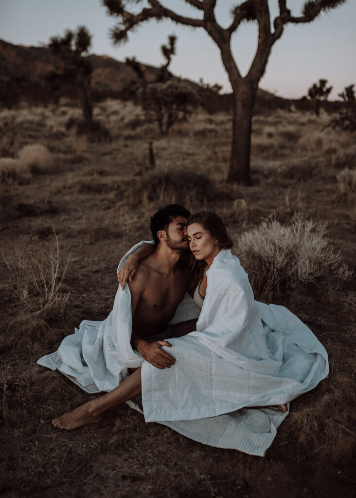 A man kissing a woman on the cheek. They are in Joshua Tree and sitting on the ground in their underwear covered with a comforter during their couples boudoir photoshoot