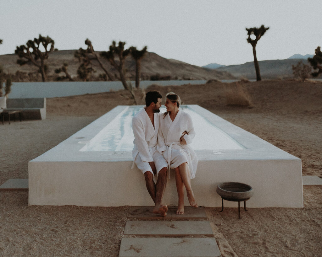 A wedding couple in white bathrobes sitting on the edge of the pool at their airbnb in Joshua Tree.
