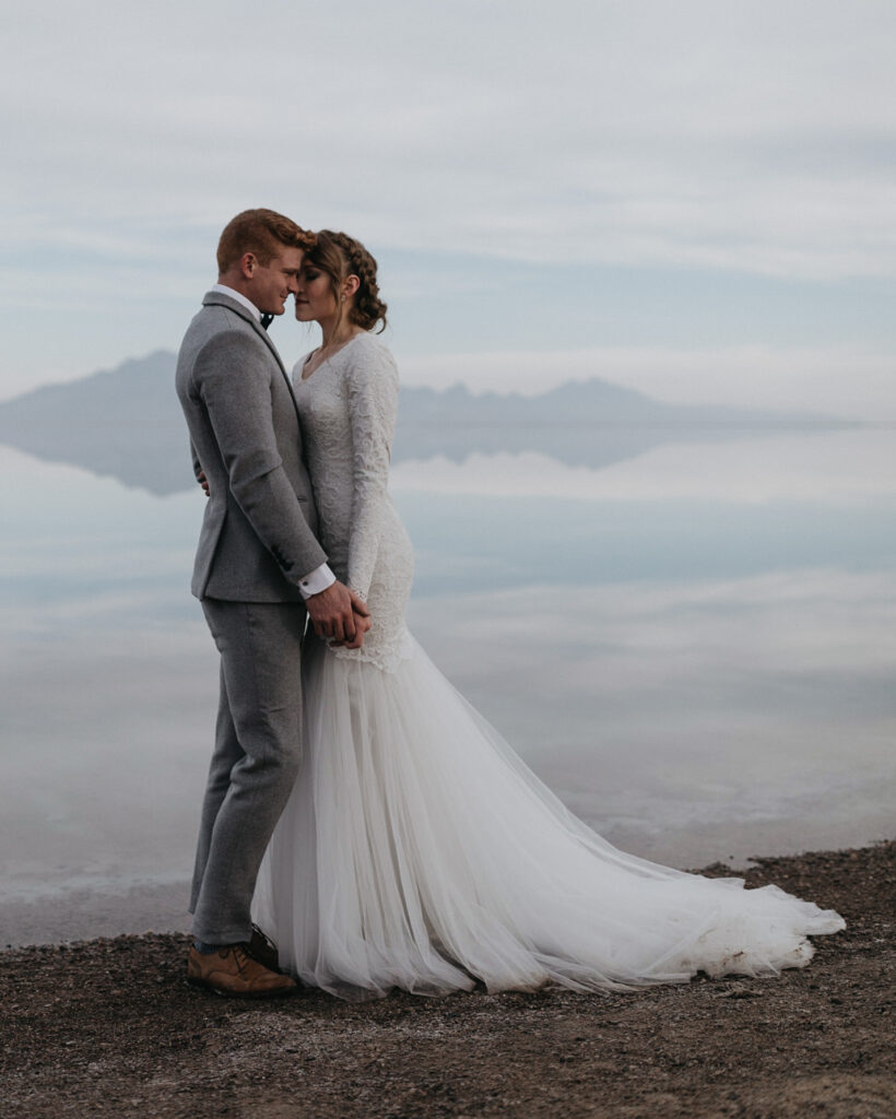 A luxury elopement couple pressed against each other with the Bonneville Salt Flats in the background, covered in water mirroring the mountains in the distance.