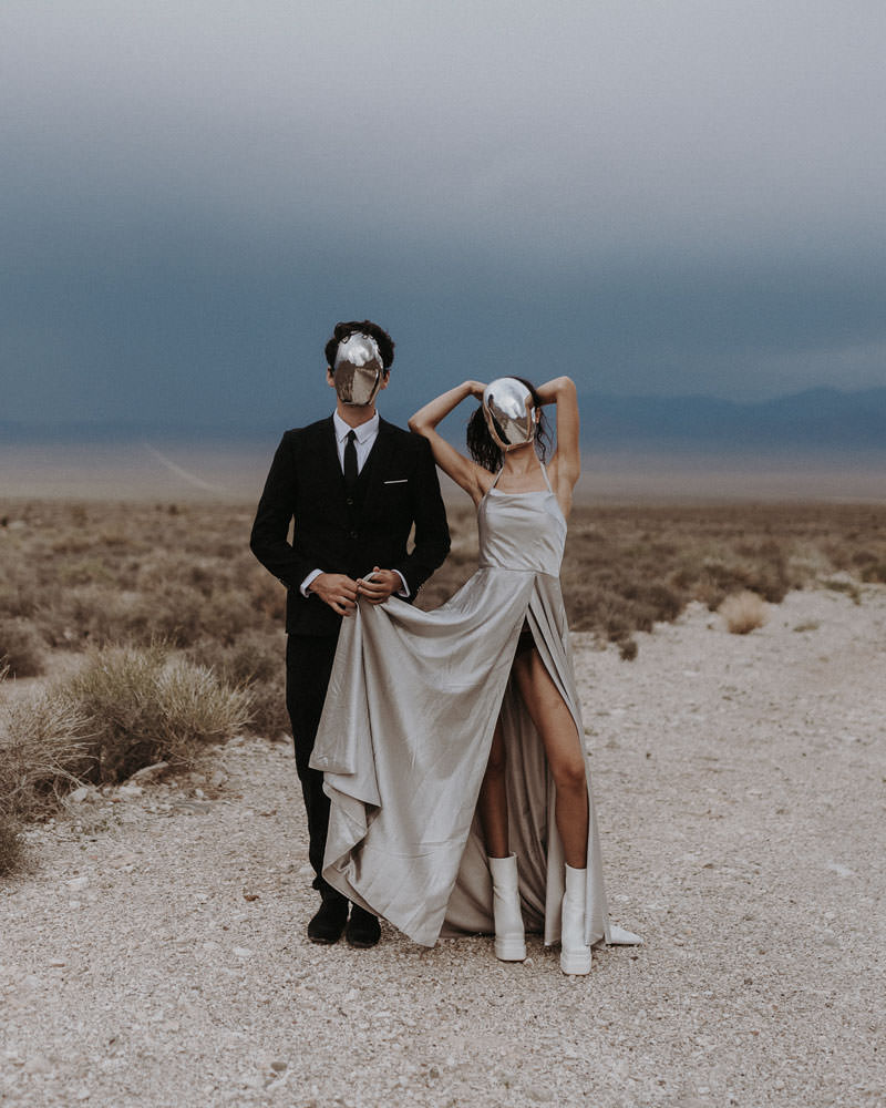 An unconventional couple having a sci-fi themed unique elopement outside of Area 51 in Nevada. They are wearing mirror masks and the bride is wearing a silver metallic dress.