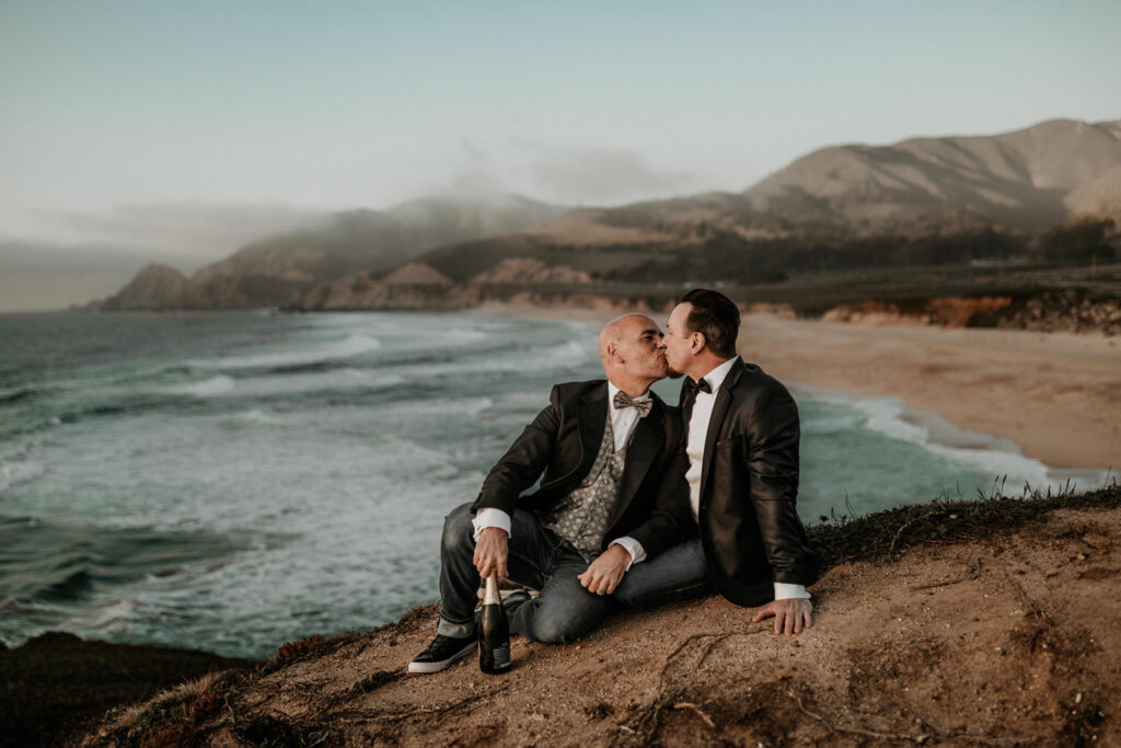 A gay elopement couple kissing on the shores of Big Sur with a bottle of champagne.