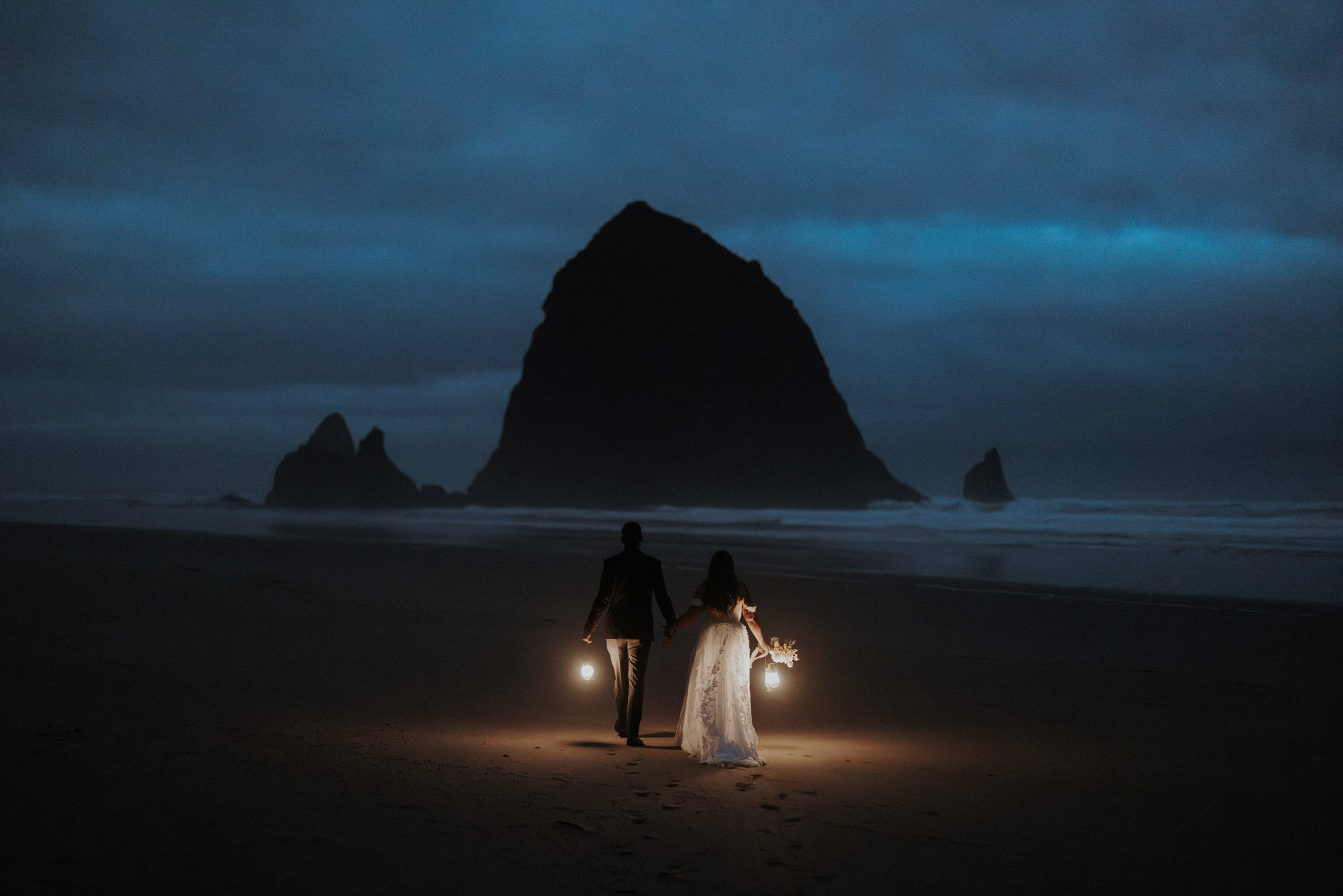 An elopement couple with lanterns at Cannon Beach before sunrise, with Haystack Rock in the background as they walk away from the camera towards it.